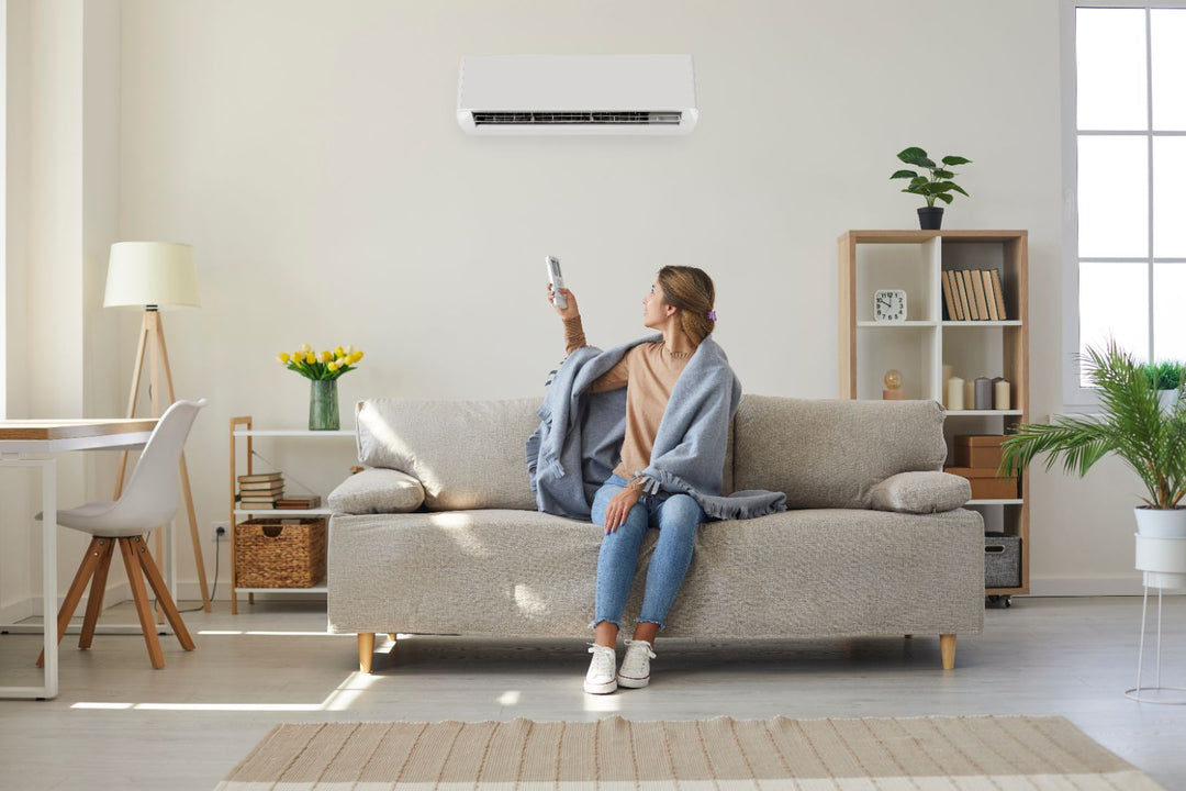 A woman sitting on a couch in her home using a remote control to adjust the settings on a mini split behind her.