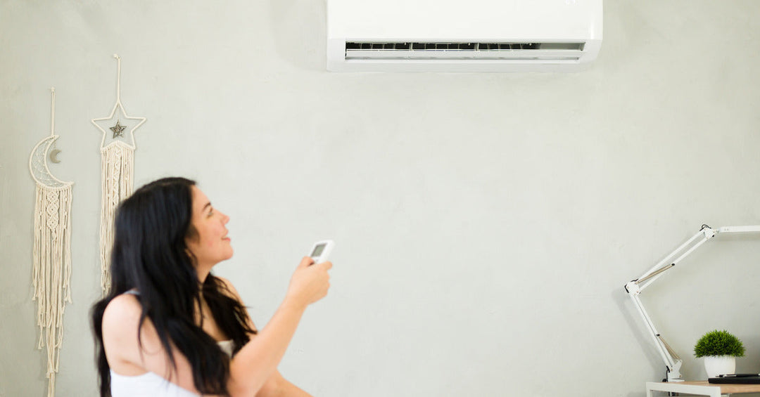 A woman smiles as she sits and points a remote control toward a mini split system to adjust the temperature in the room.