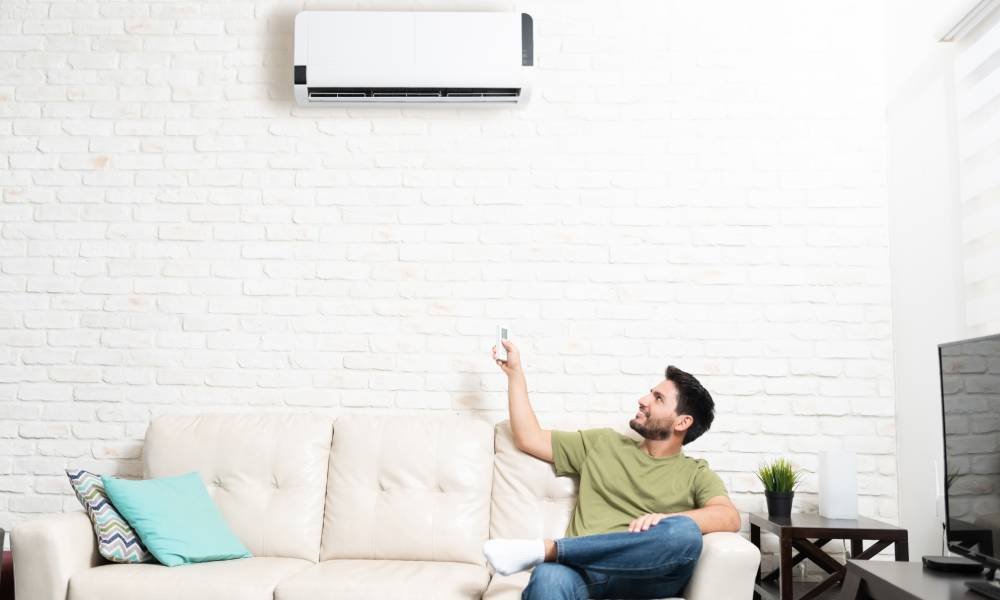 Happy man looking up at his mini split and using a remote control to adjust the temperature. He is sitting on a white couch against a white brick wall.