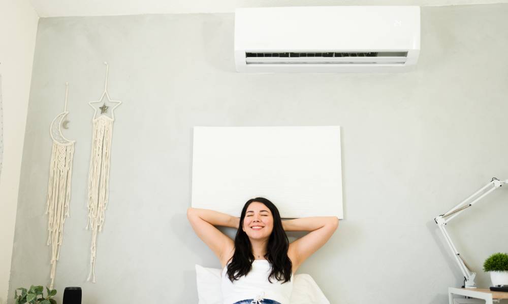 Woman in shorts and a t-shirt happily stretches out in bed under a mini split unit blowing cool air.