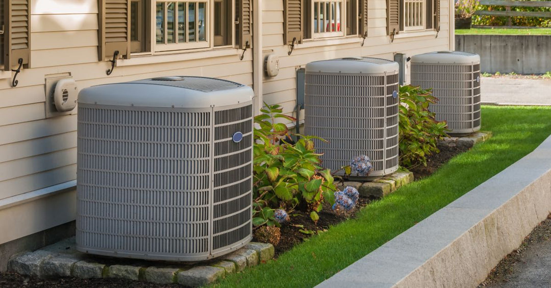 Three grey air conditioning condensers sit outside a series of homes on a slim stretch of grass by a paved road.