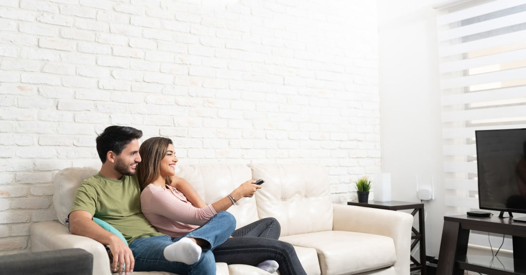 A man and woman sit on a couch watching television in front of a white brick wall topped by a mini split unit.