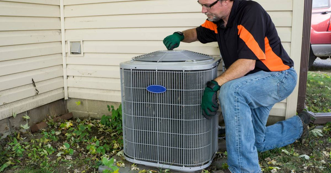 A repairman wearing glasses and gloves works on an outdoor air conditioning condenser unit set up beside a house.