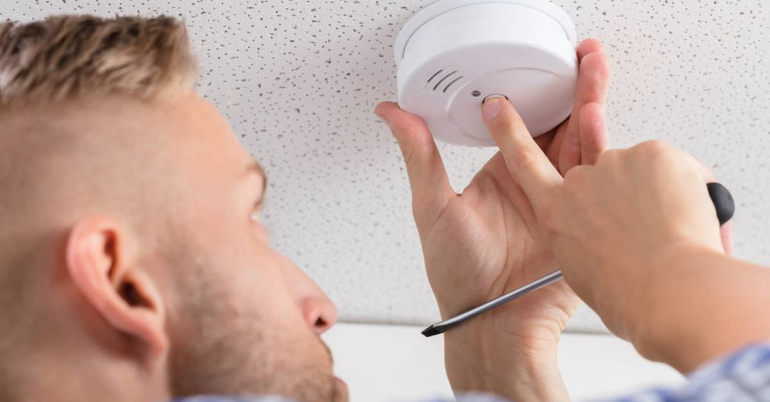 A man holding a screwdriver and installing a CO detector on the ceiling. He is pressing a button on the device.