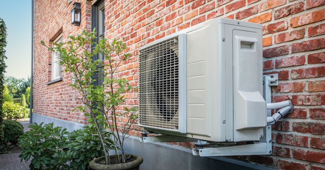 A white metal heat pump device is sitting on the side of a red-bricked house. Several plants are nearby.